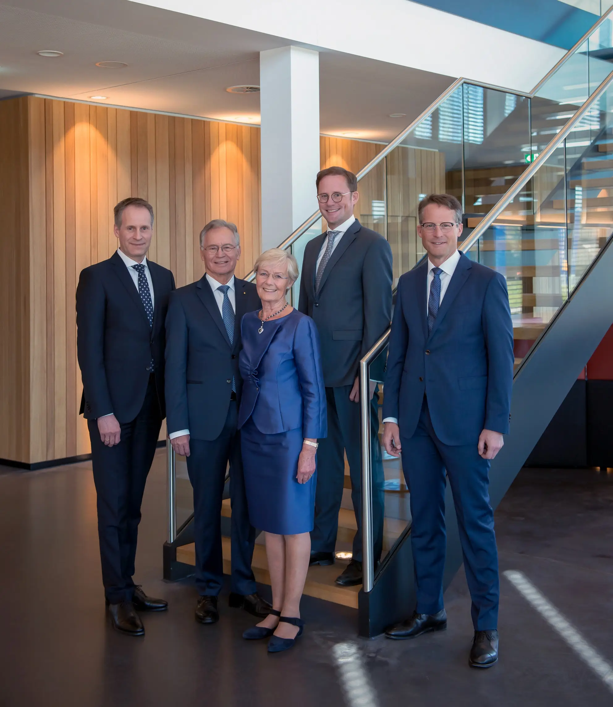 Fünf Personen in Anzügen und businessgerechter Kleidung stehen in einem modernen Bürogebäude vor einer Treppe.